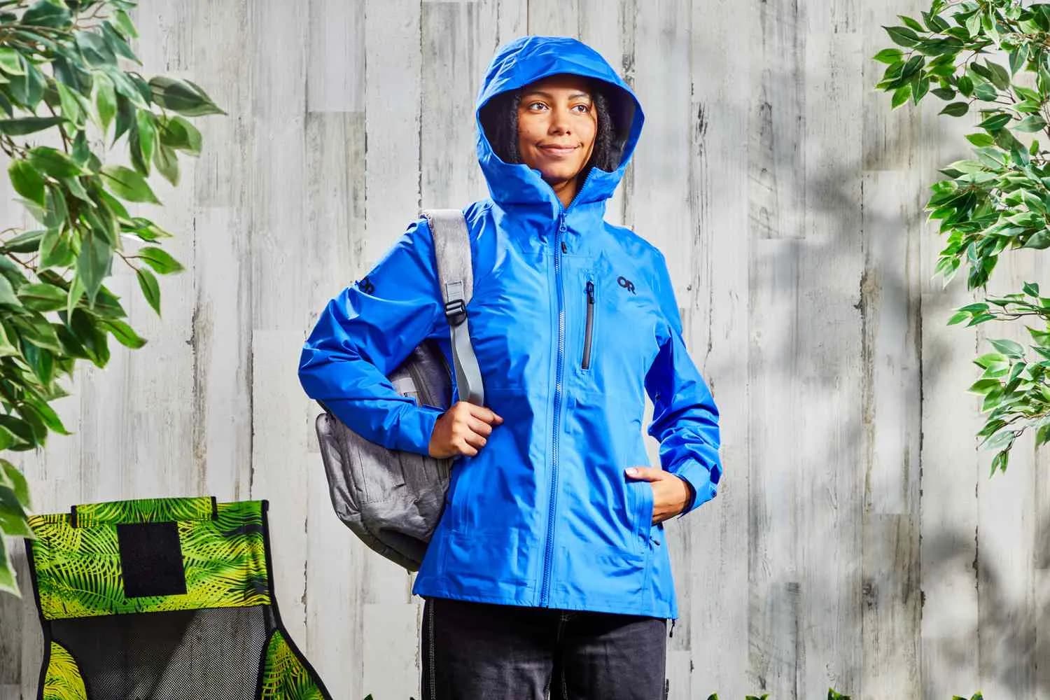 A woman wearing a lightweight jacket and a stylish hat stands confidently outdoors, showcasing a practical yet fashionable look against a vibrant natural backdrop