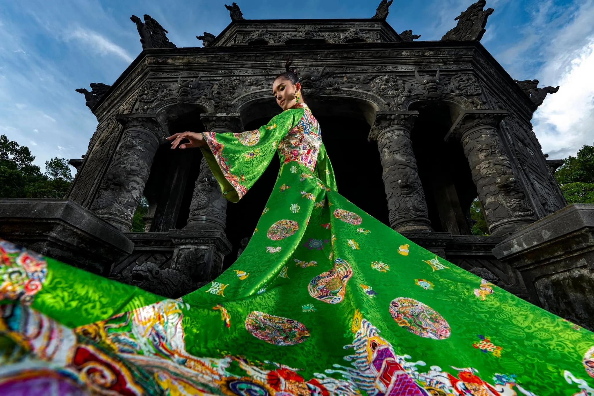 Model in a hand-embroidered silk áo dài with traditional motifs by designer Trần Thiện Khánh, standing at the ornate Khải Định Tomb in Huế, showcasing the vibrant fusion of fashion and Vietnamese heritage