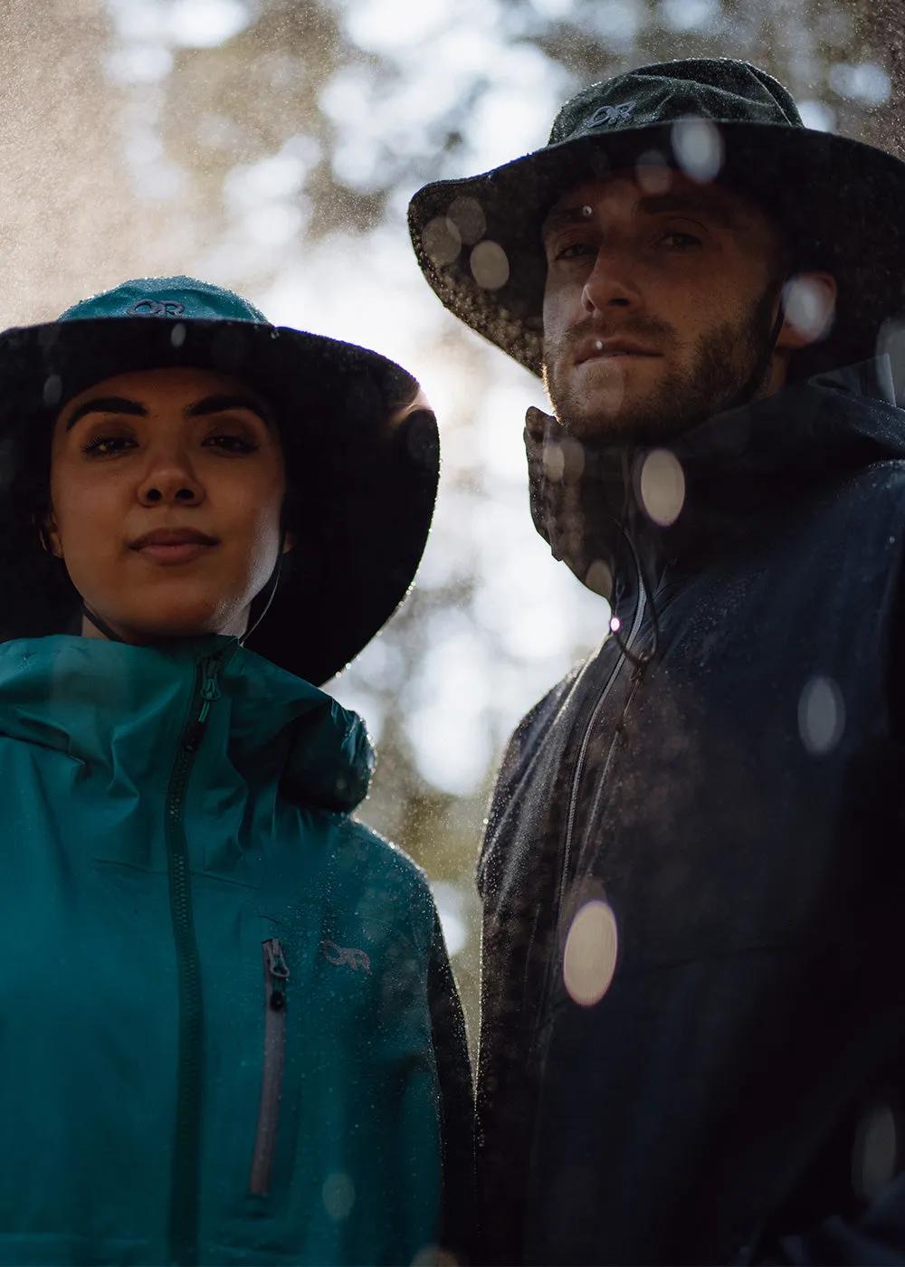 A couple wearing matching rain jackets and hats stands outdoors on a rainy day, smiling at each other as raindrops fall on their jackets