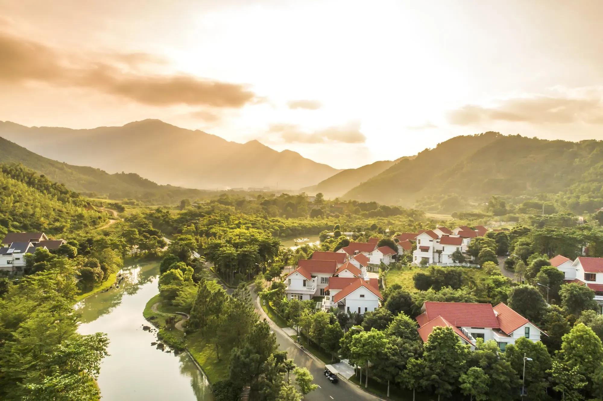 A residential area nestled in lush nature, with sunlight streaming through clouds over the hills.