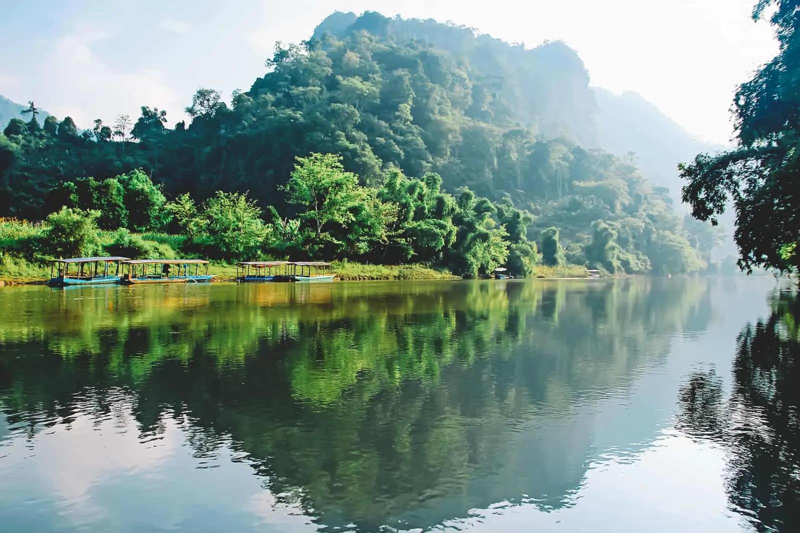A serene view of Ba Be Lake on a sunny day, with clear blue skies reflecting on the calm waters, surrounded by lush mountains and greenery in Ba Be National Park.