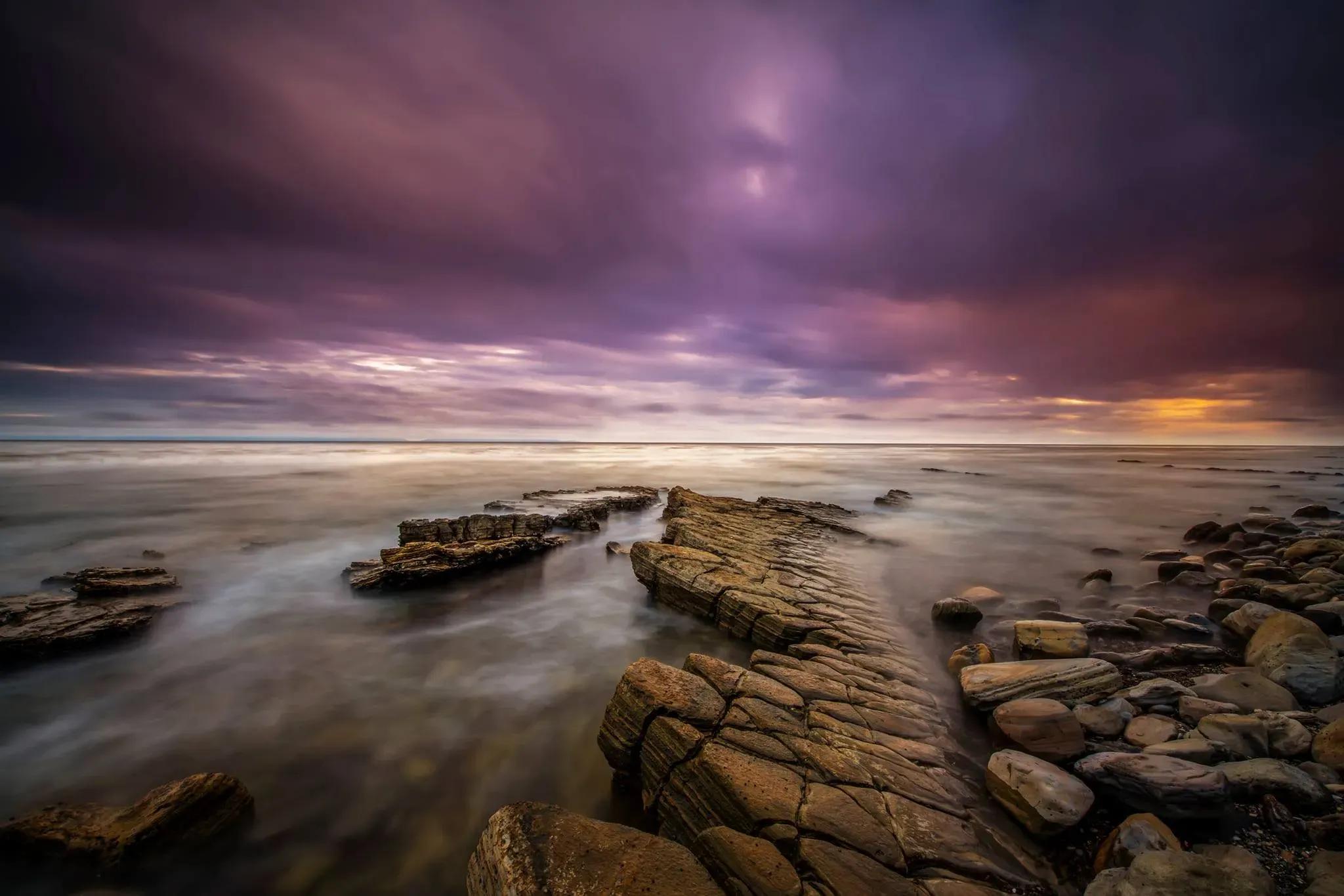 A stunning skyline with shades of purple reflecting over the ocean, with rocky coastline in the foreground
