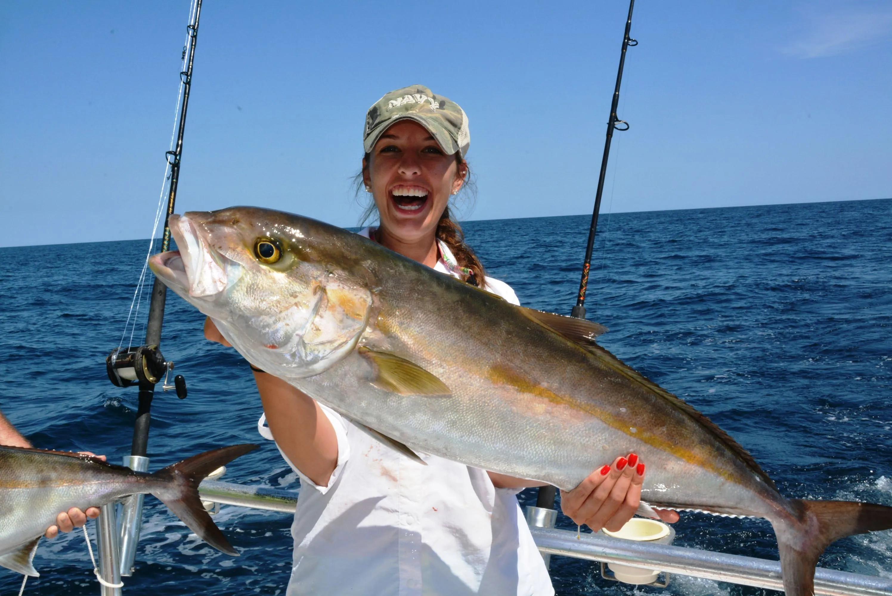 A woman participating in a sea sport fishing event holds up a large herring she just caught, smiling proudly as she stands on a boat with the ocean in the background