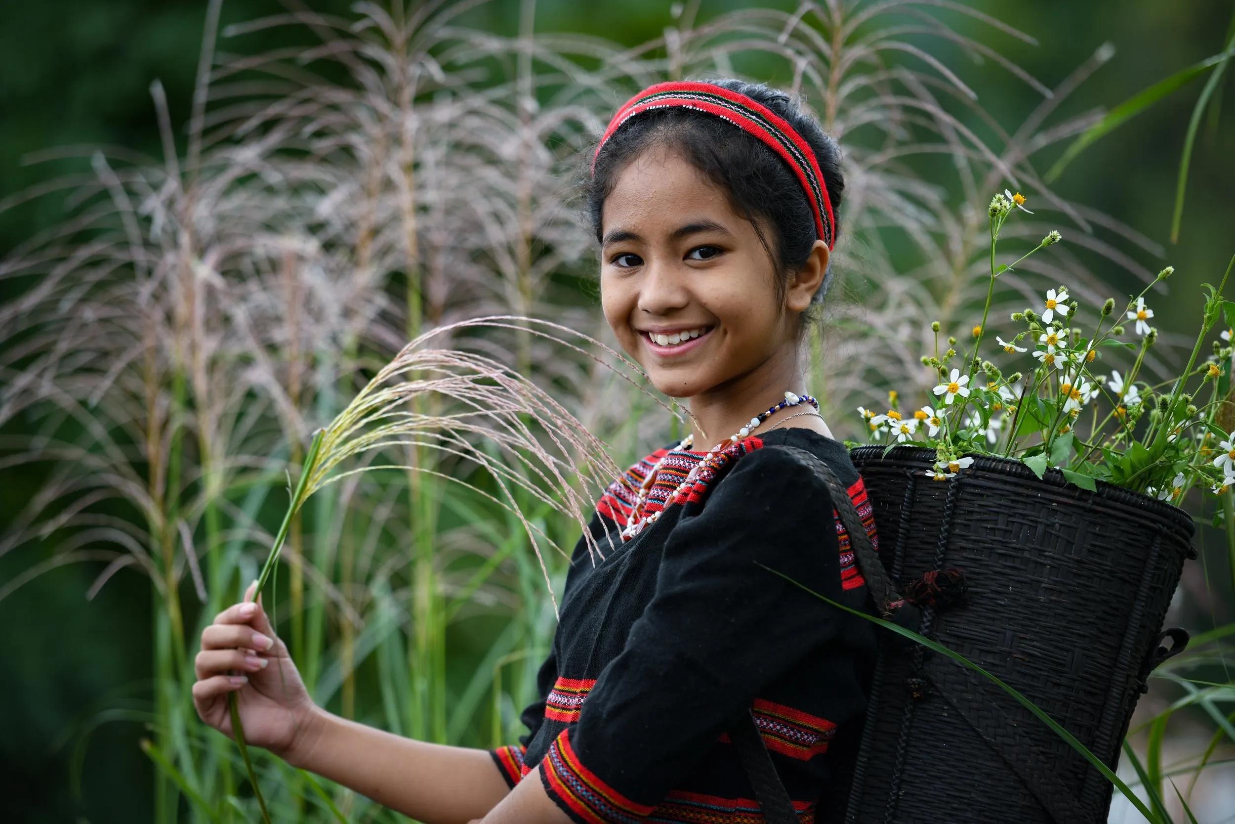 Tà Ôi girl in traditional attire with a vibrant flower basket, smiling brightly amidst vast nature.