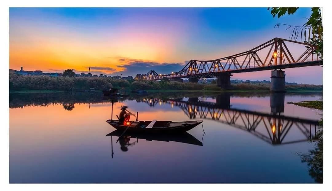 The Long Bien Bridge bathed in the warm glow of sunset, with an old fisherman sitting on a small boat casting his net on the calm river below