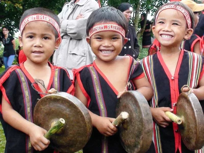 Three joyful Ro Mam boys in traditional attire, wearing headbands and holding drums.