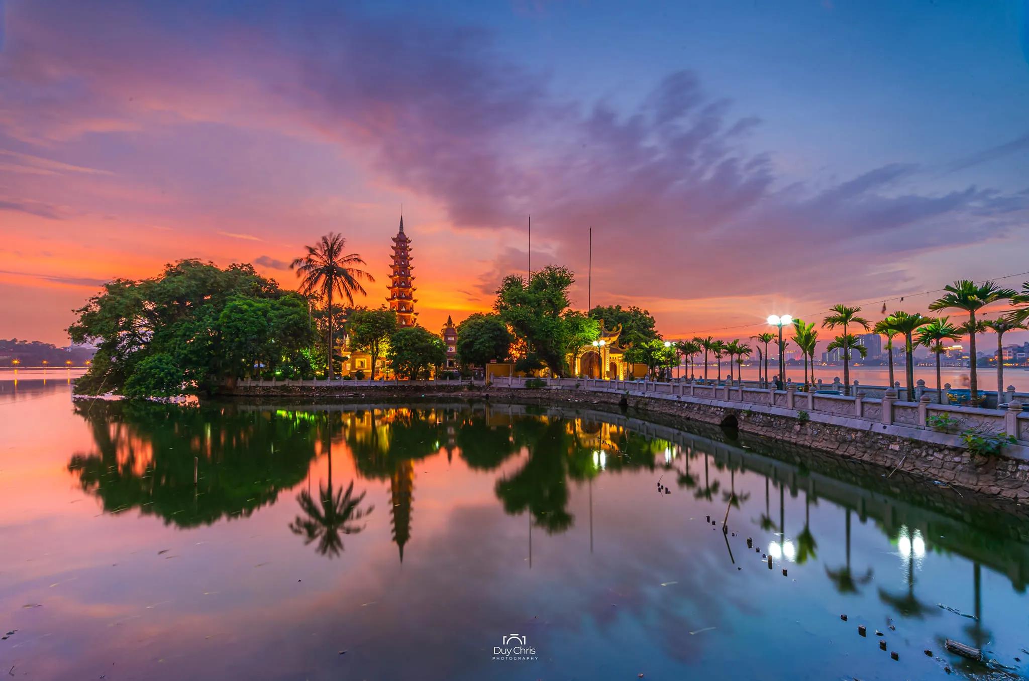Trấn Quốc Pagoda at sunset, its golden reflection shimmering on the calm waters of West Lake, surrounded by lush greenery