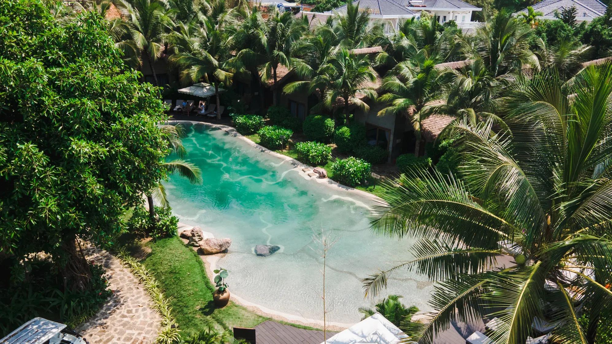 The photo captures a refreshing green swimming pool surrounded by lush trees, offering maximum relaxation for guests at M Village Phú Quốc.