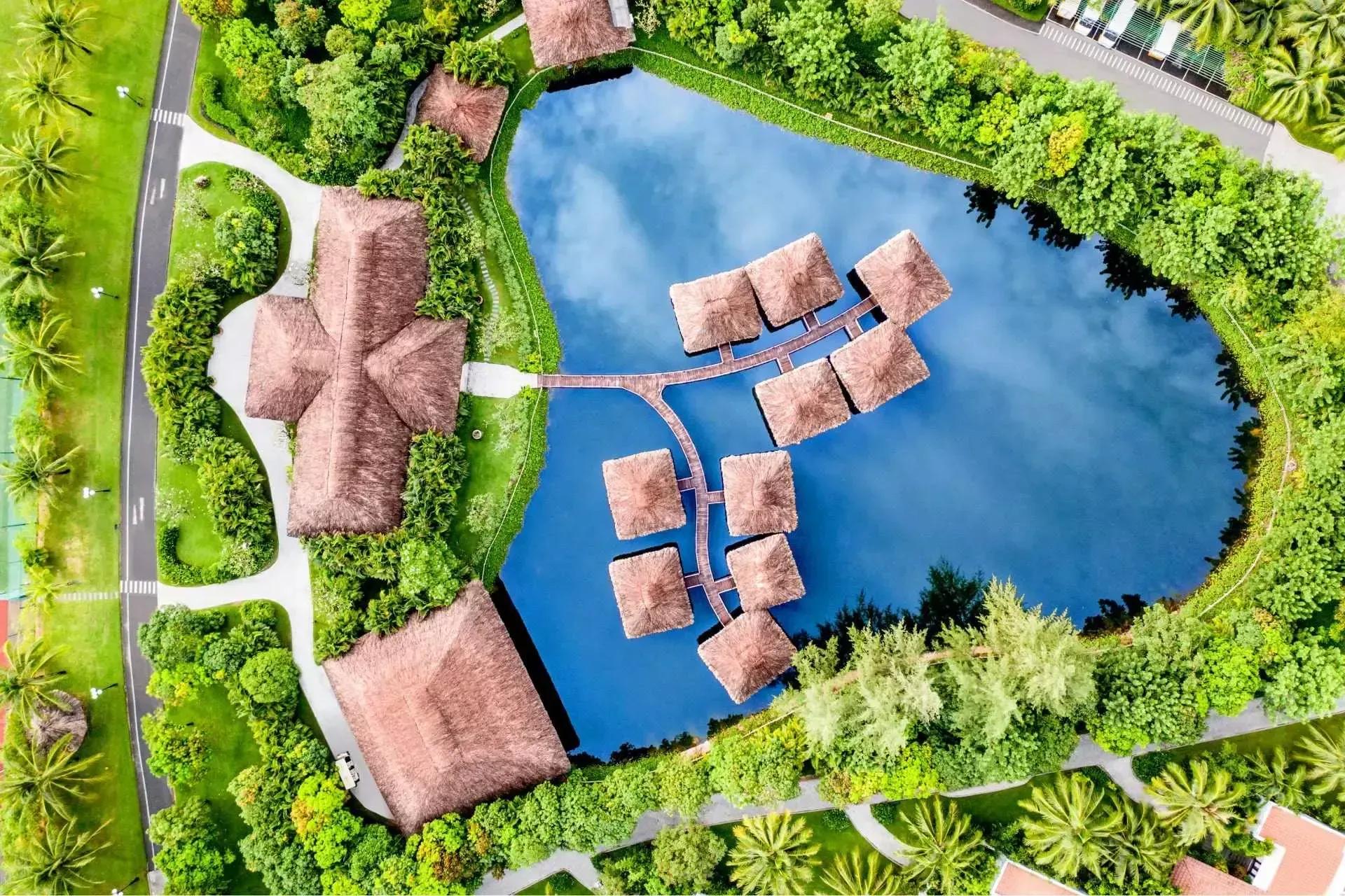 Resort area with thatched roofs and a blue lake viewed from above, surrounded by greenery and walking paths.